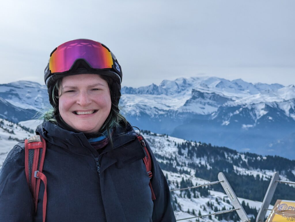 A trans woman wearing a skiing jacket, ski helmet and goggles stands in front of a mountain vista