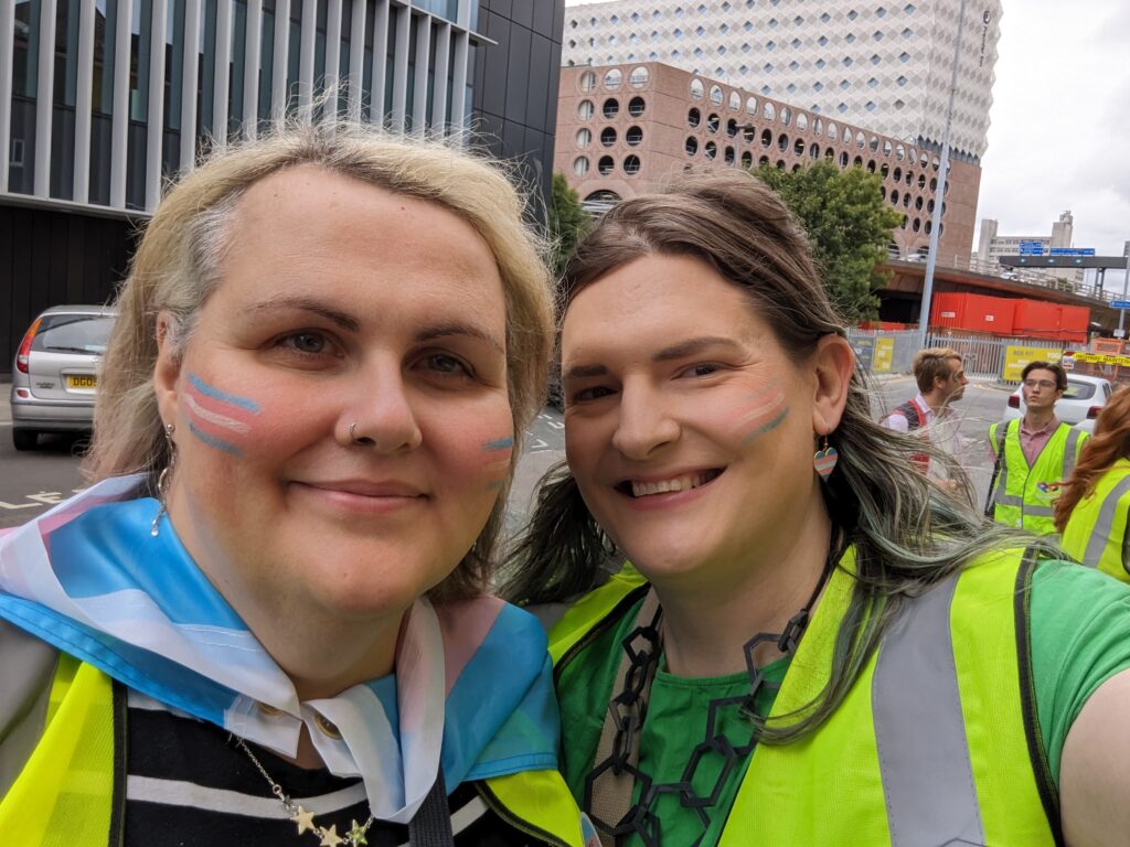 Two trans women taking a selfie, both are in hi-vis and have trans face paint on