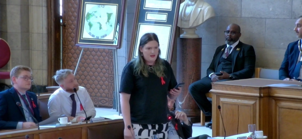 Chris Northwood, wearing a red ribbon, stands in the council chamber and reads from her phone