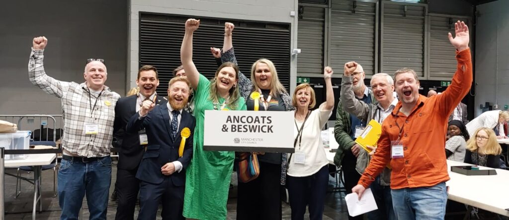 A group of people celebrating, wearing Lib Dem rosettes and holding a sign saying "Ancoats and Beswick"