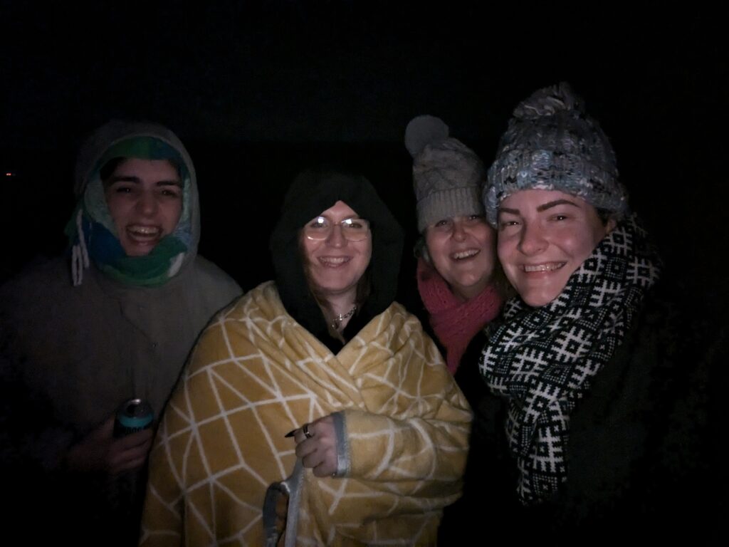 Four trans girls, lit only by flash on a dark night, looking into a camera wrapped up in warm clothes and blankets.