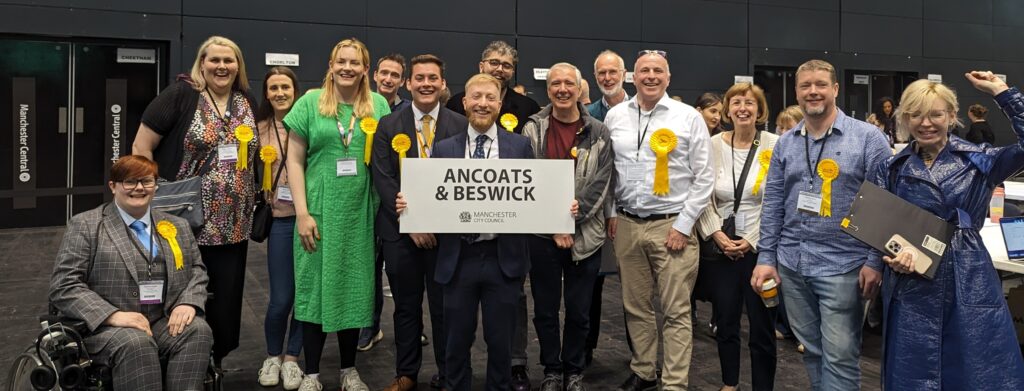 A group of people wearing yellow Lib Dem rosettes look happy behind Alan Good holding a sign that says Ancoats & Beswick.