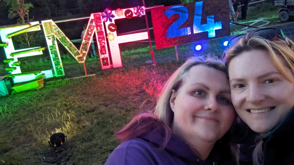 Chris and Meg stand in front of a sign saying EMF 24, in a field lit up by colourful lights.