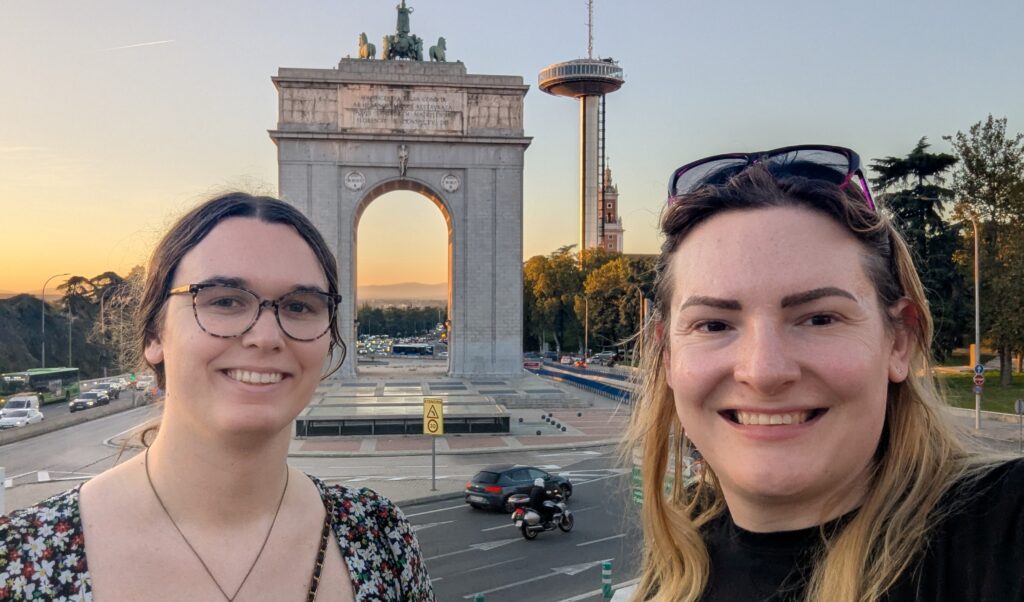 Chris and Emily stand in front of an arch and tower in Madrid, looking into the camera.