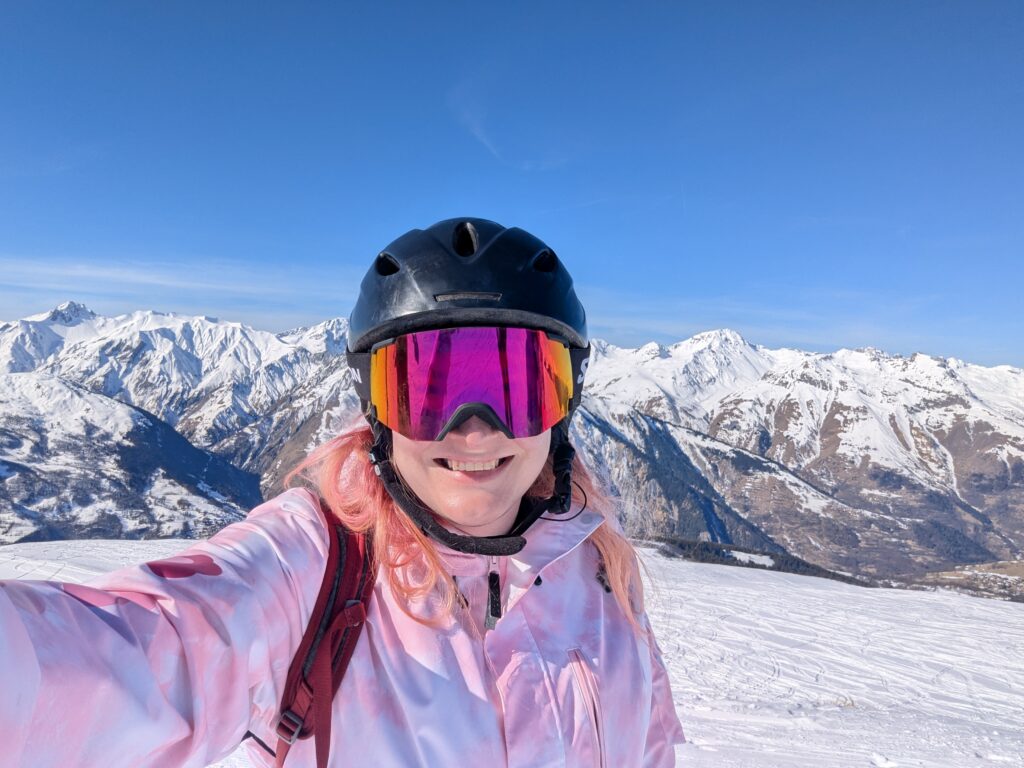 Chris takes a selfie smiling into the camera wearing ski gear, on a mountain with a mountain backdrop