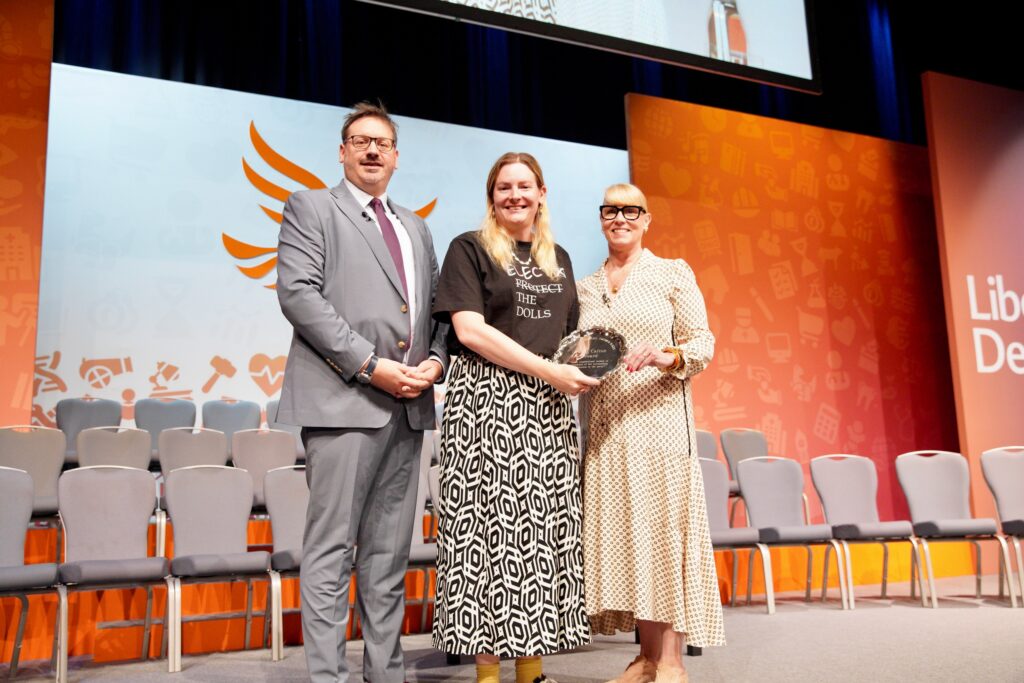 Chris Northwood stands on stage at Lib Dem conference holding the Patsy Calton award stood next to FCC chair Nick da Costa and Lib Dem Women chair Donna Harris
